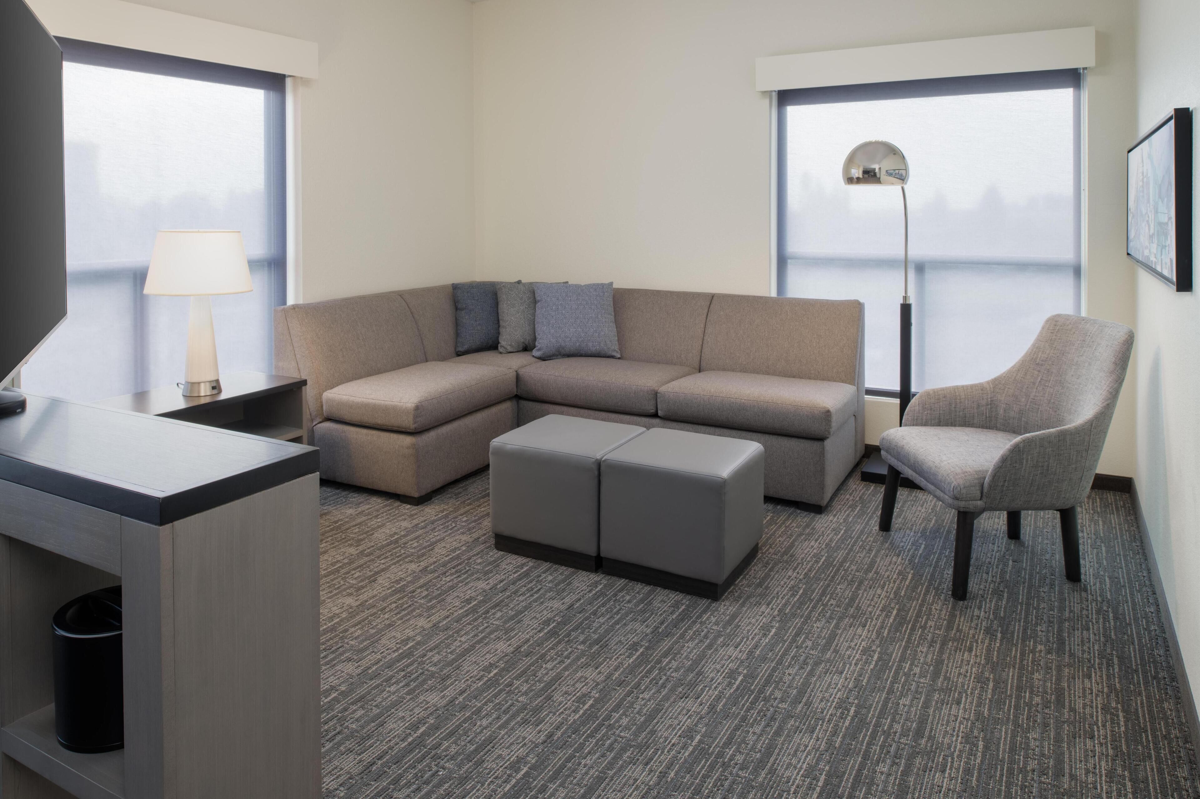 Una habitación de hotel moderna con sofá modular, un pequeño reposapiés, una silla, un escritorio con lámpara y ventanas amplias con vistas.