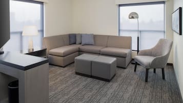 Una habitación de hotel moderna con sofá modular, un pequeño reposapiés, una silla, un escritorio con lámpara y ventanas amplias con vistas.