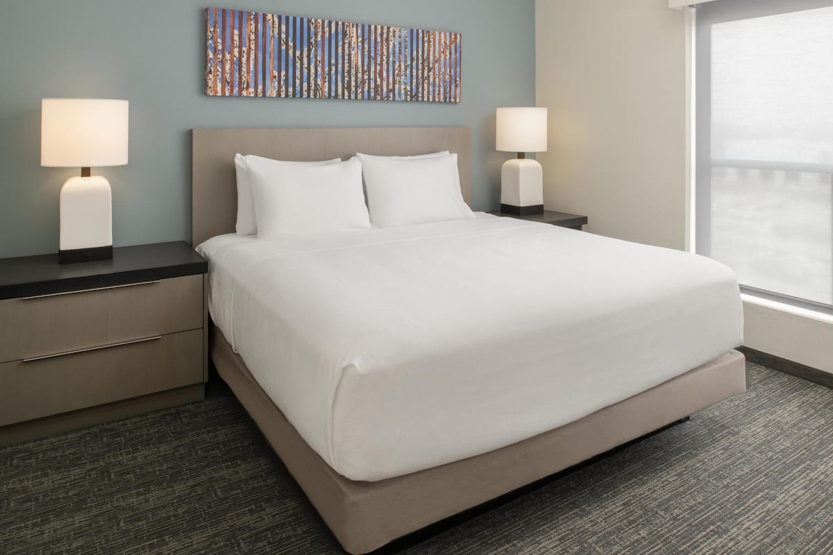 A hotel room with a large bed, two bedside tables with lamps, a headboard, a window, and a colorful abstract painting on the wall.