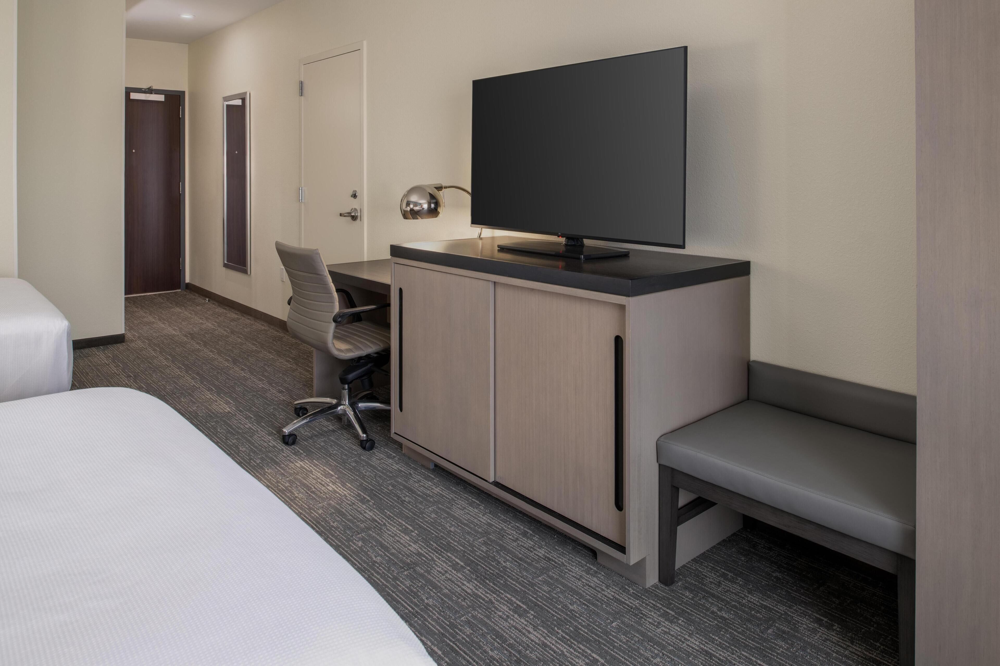 A hotel room with a bed, a desk with a chair, a television, and a bench.