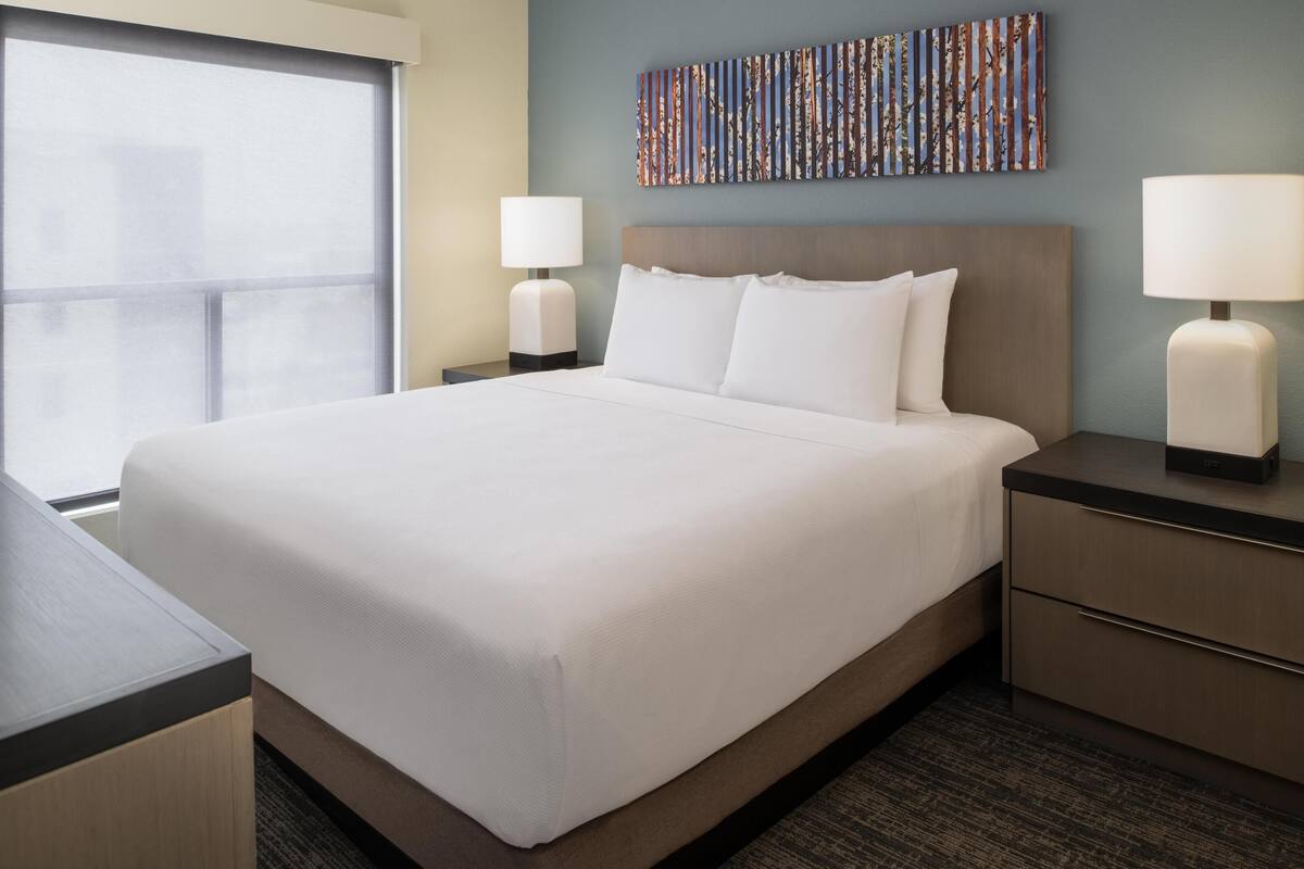 A hotel room with a large bed, two bedside tables with lamps, a wall art piece, and a window.