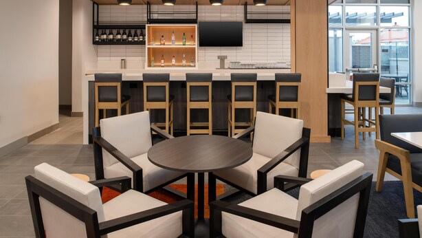 A modern bar area with a round table and chairs, a shelf with bottles, and a flat-screen TV. - Hyatt House Fresno Campus Pointe (Fresno)