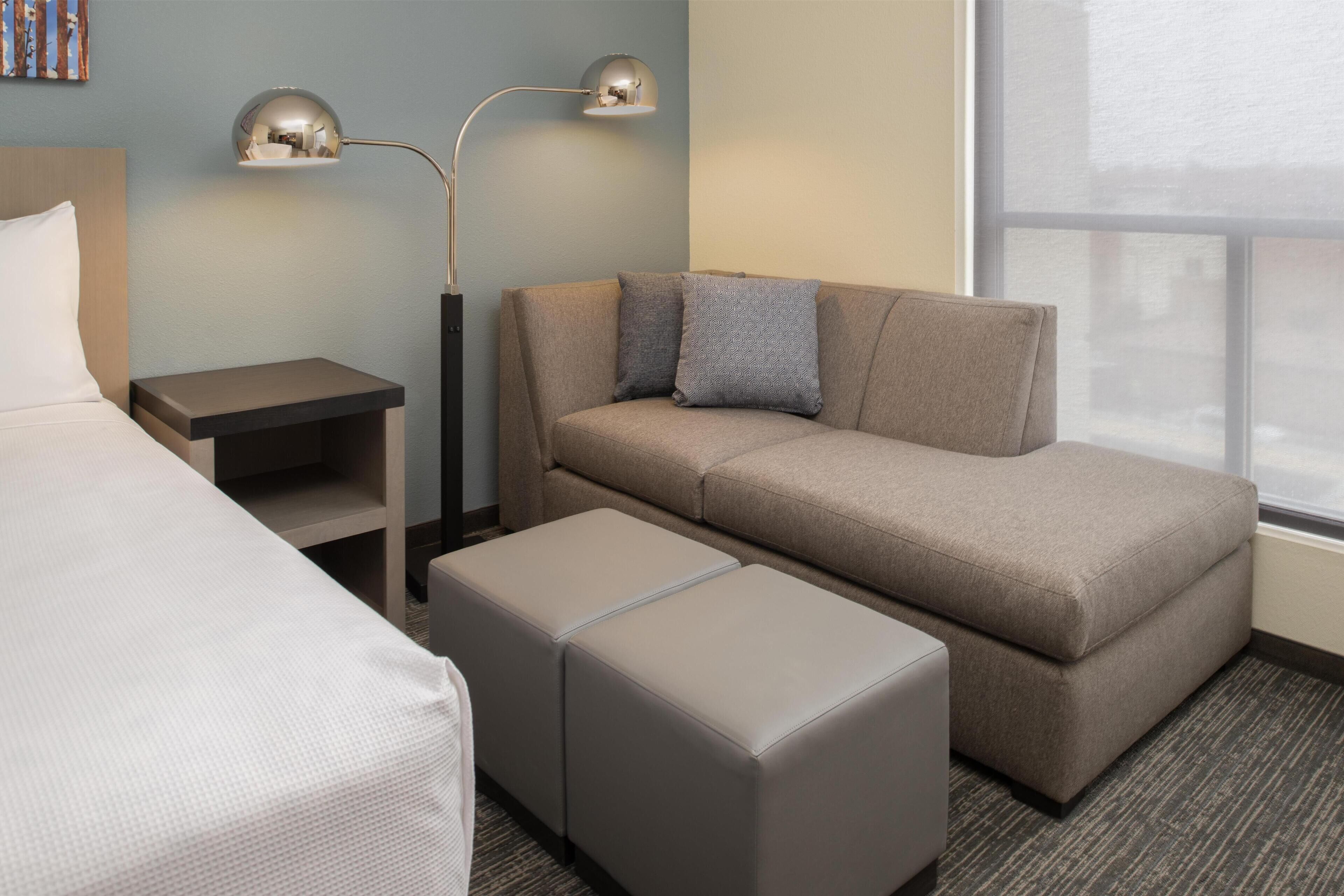 A hotel room with a bed, a sofa, two ottomans, a nightstand, and a lamp.
