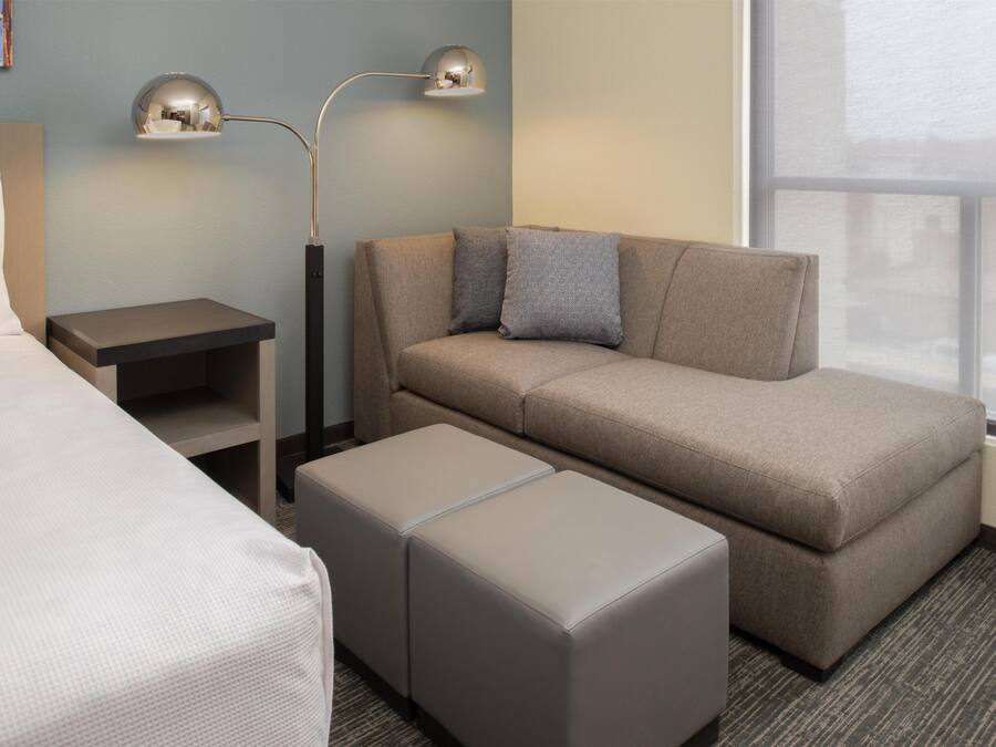 Quarto de hotel com cama, sofá, dois pufes, mesa de cabeceira e abajur.