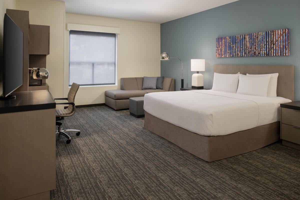A hotel room with a large bed, a desk with a computer, a chair, a sofa, and a window with blinds.
