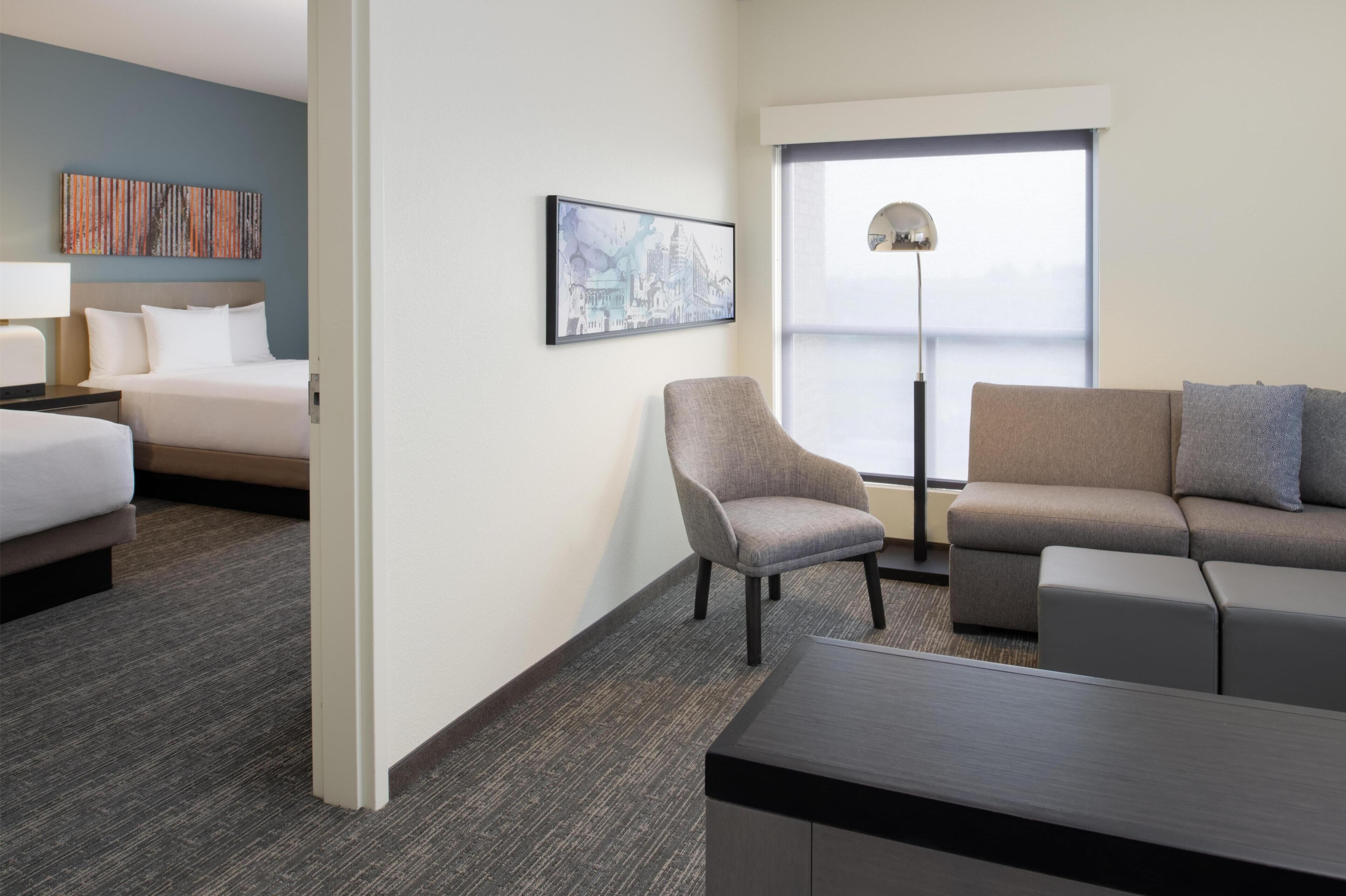 Ein Hotelzimmer mit einem Bett, einem grauen Sessel, einem grauen Sofa, einem kleinen Tisch und einem großen Fenster mit Blick.
