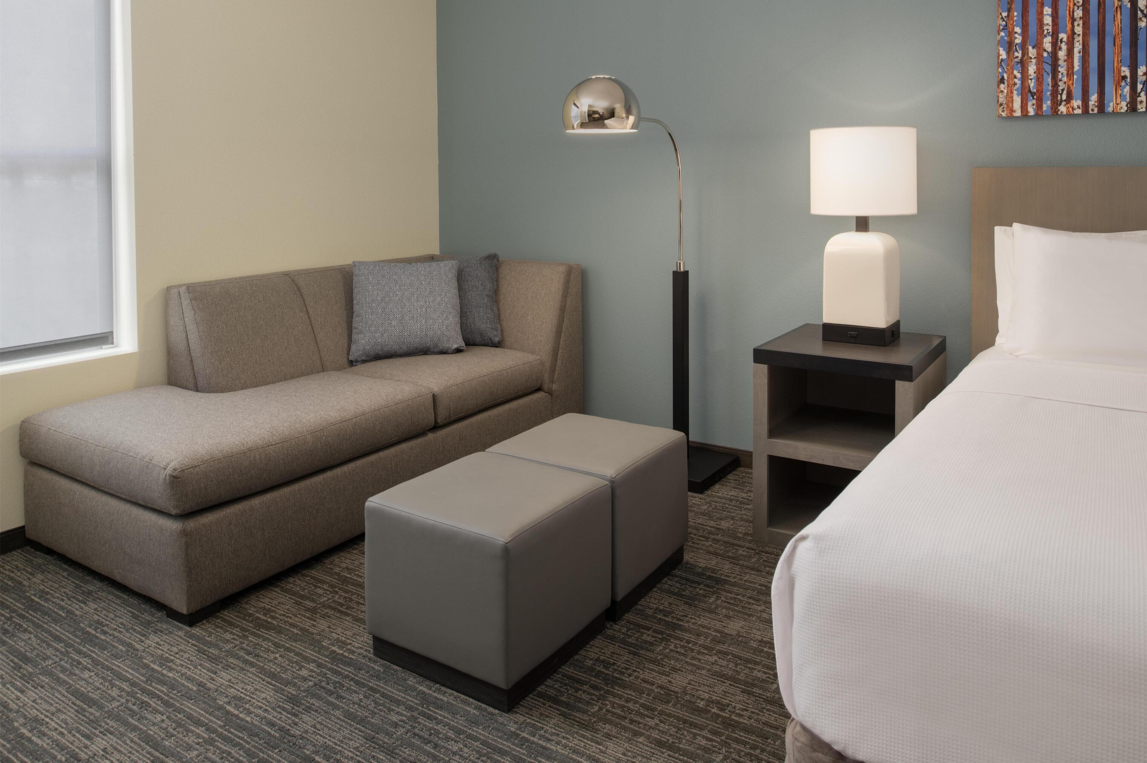 A hotel room with a sofa, a small ottoman, a nightstand with a lamp, and a bed.