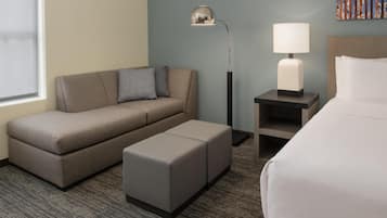 A hotel room with a sofa, a small ottoman, a nightstand with a lamp, and a bed.