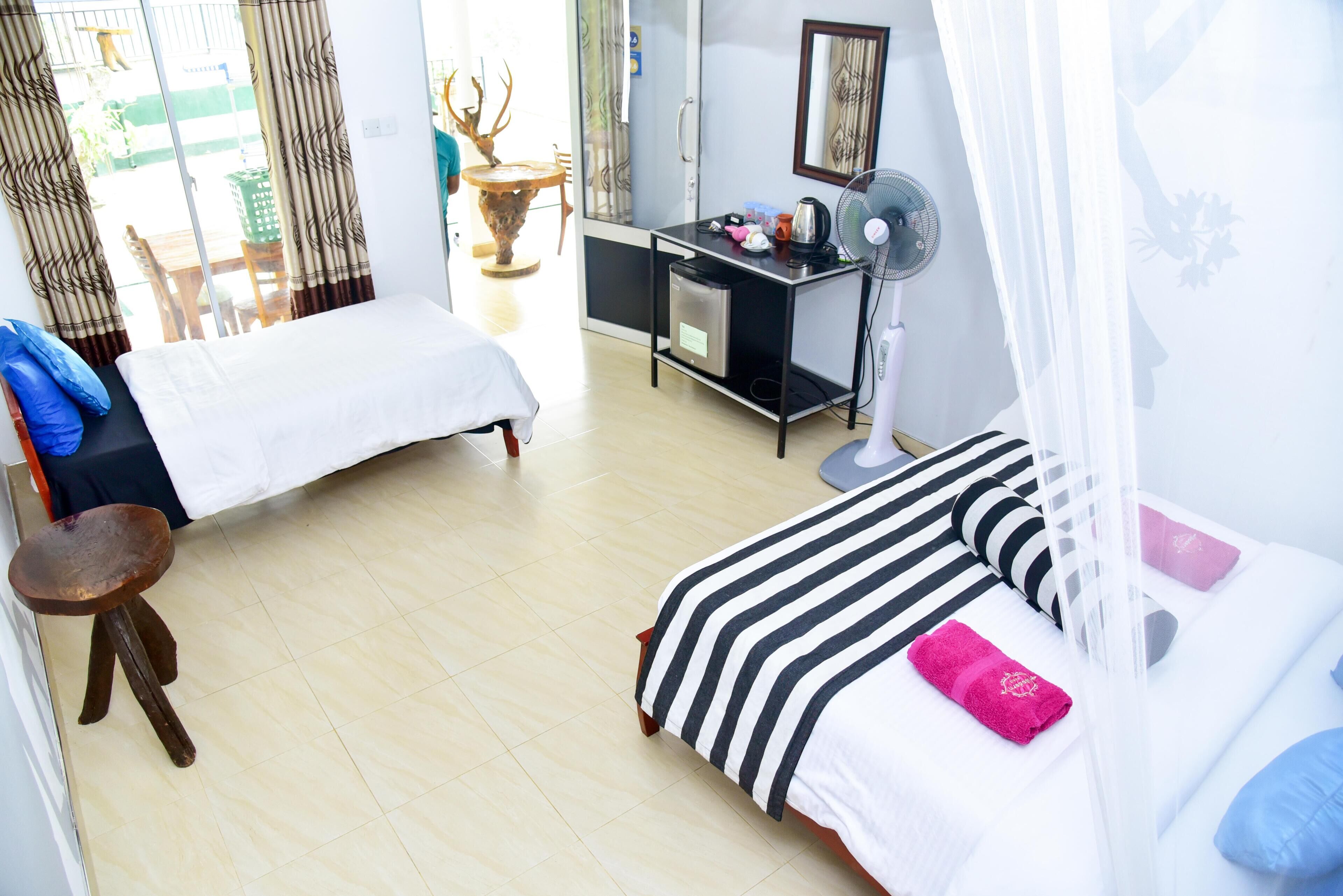 A hotel room with two beds, a desk with a fan, a small stool, and a view of the outside.