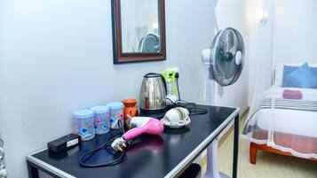 A room with a bed, a bedside table with a fan, a kettle, and a hairdryer.