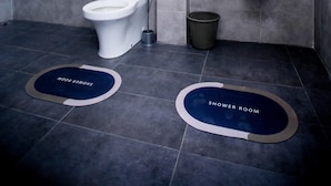 Un baño moderno con inodoro, lavabo y ducha. Hay dos alfombras en el suelo con la inscripción 'Sala de Duchas'.