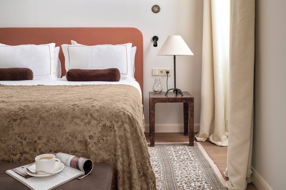 Una cama bien hecha con una colcha estampada, una mesita de noche con una lámpara y una revista enrollada sobre una pequeña mesa.
