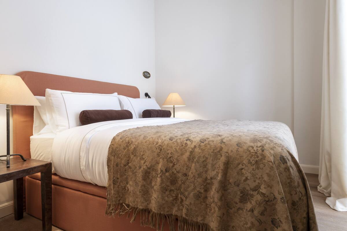 Una cama bien hecha con un edredón estampado, dos lámparas de noche y una mesita con lámpara.