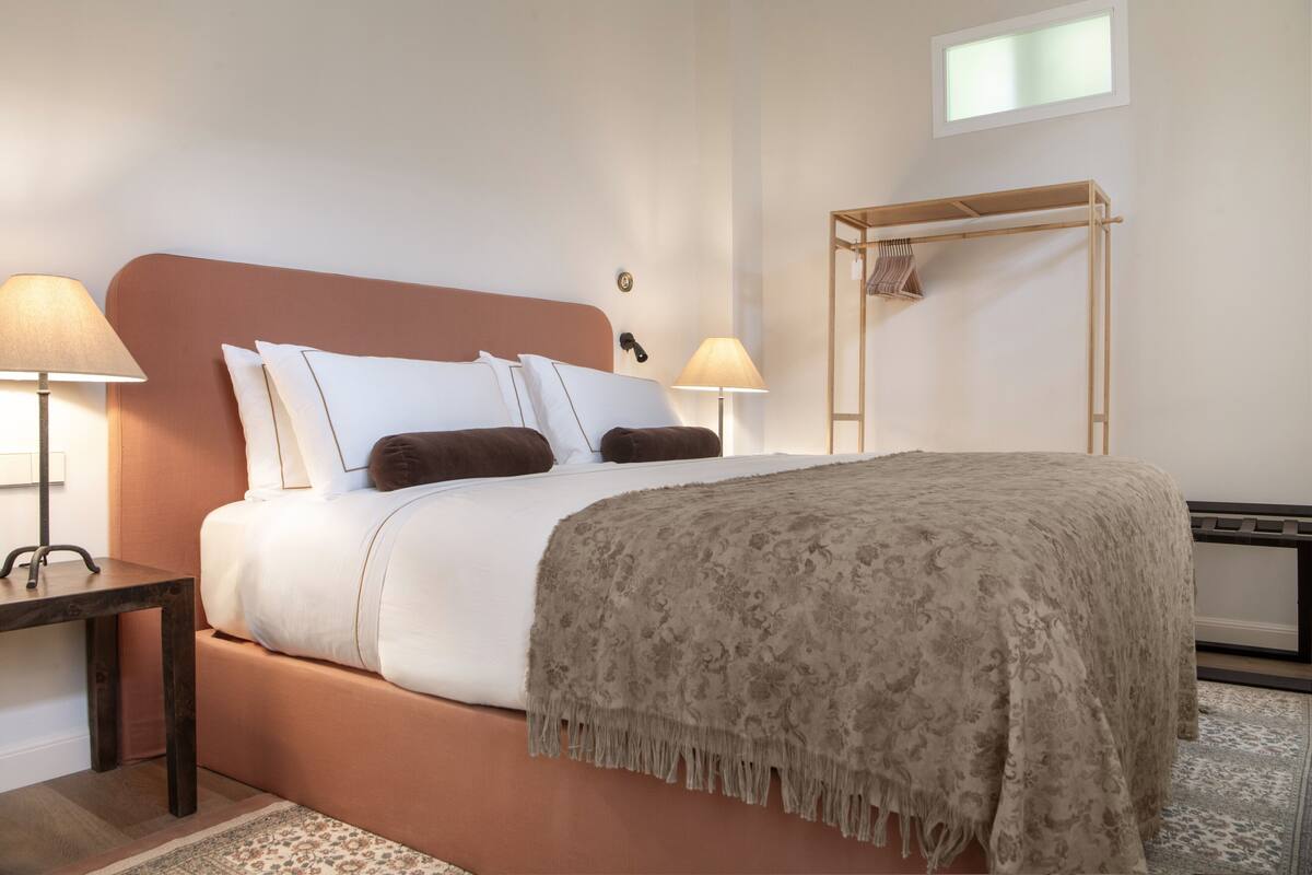 Un dormitorio ordenado con cama, dos mesitas de noche, lámparas, un perchero de madera y un ventanal.
