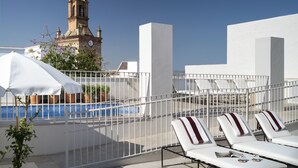 Een dakzwembad met ligbedden, een witte parasol en een klokkentoren op de achtergrond.