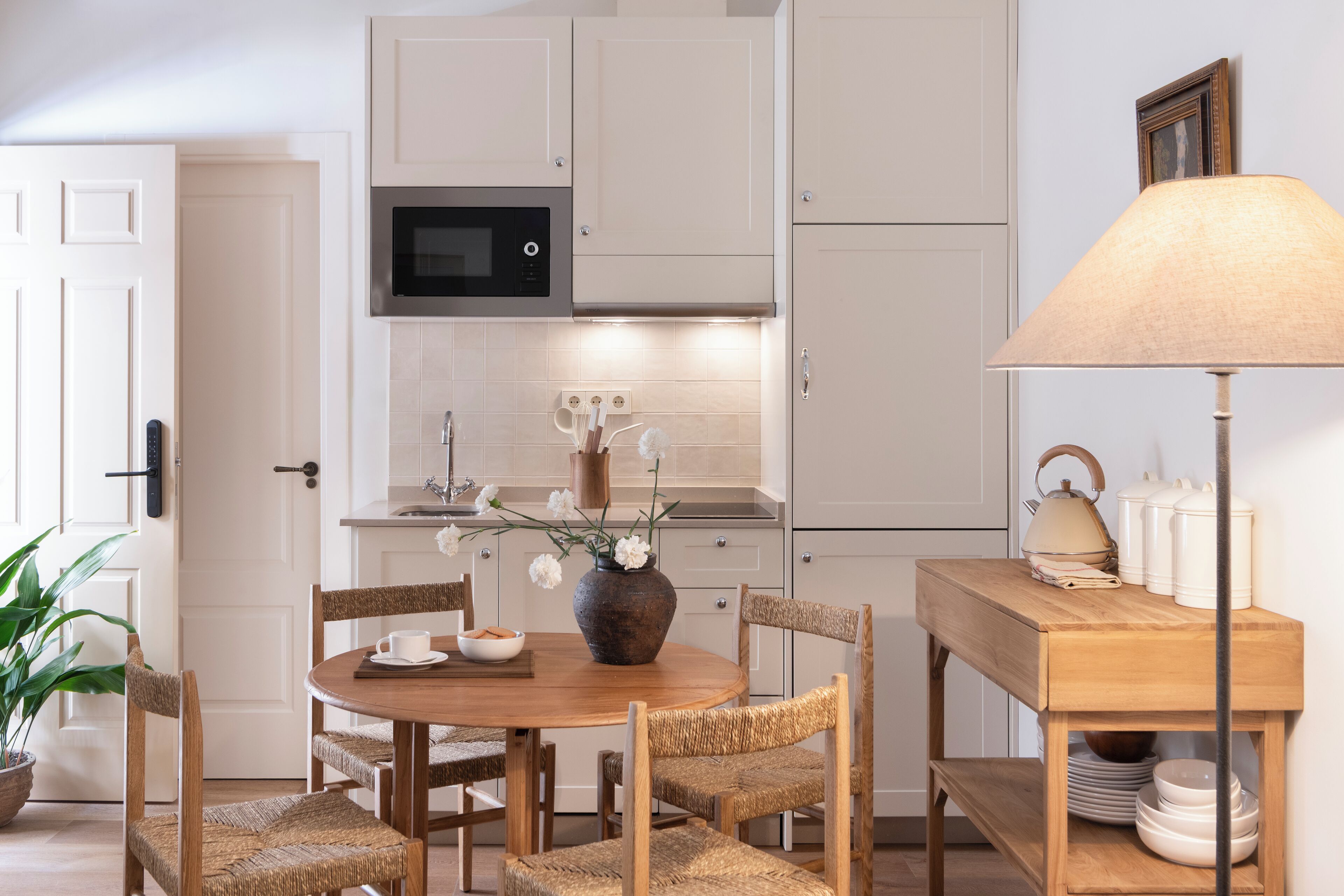A modern kitchen with white cabinets, a microwave, a wooden dining table with chairs, a vase with flowers, and a lamp.