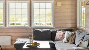 A cozy room with a grey sofa, a small table with a candle and a vase, and two windows with a view of a deck. - Stavtjørn Apline lake Haven (Vikeså)