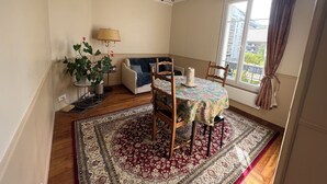 A dining area with a table set for four, a chandelier, a sofa, and a large window with curtains. - Cosy apartment (Boulogne-Billancourt)