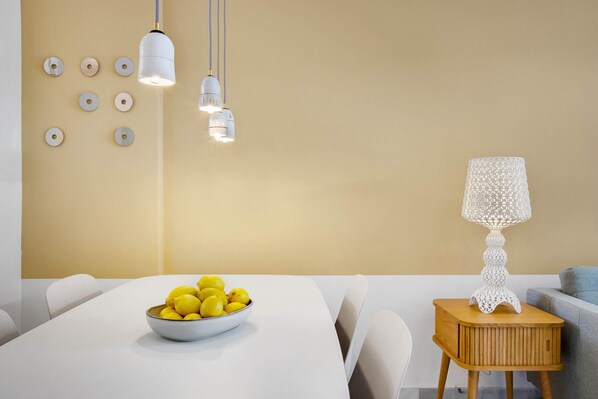 Un comedor con una mesa blanca, un cuenco de limones y una lámpara blanca sobre una mesita de madera.