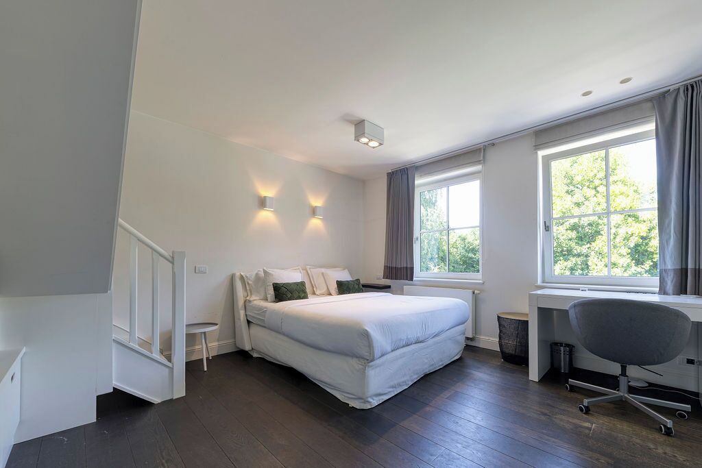 Une chambre avec un lit, un bureau et une chaise. Il y a deux fenêtres avec des rideaux, un petit tabouret et une poubelle.