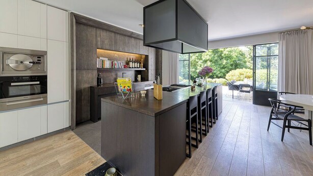 Een moderne keuken met een centraal eiland, een inbouwoven en een groot raam met uitzicht op een tuin.