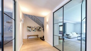Een modern interieur met een houten vloer, een trap met abstracte muurkunst, en een witte bank in een lichte kamer met glazen wanden.