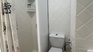 A small, compact bathroom with a toilet, a shower, and a shelf with toiletries.