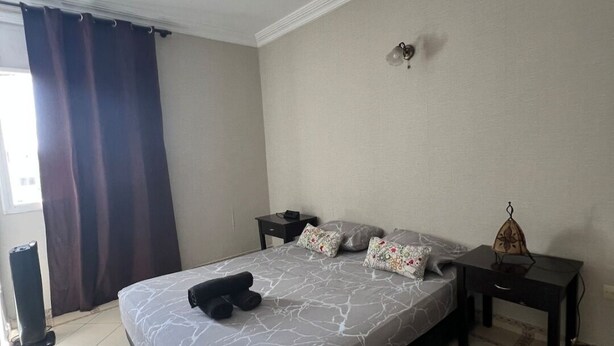 A bedroom with a bed, a nightstand, a lamp, a window with curtains, and a tiled floor.