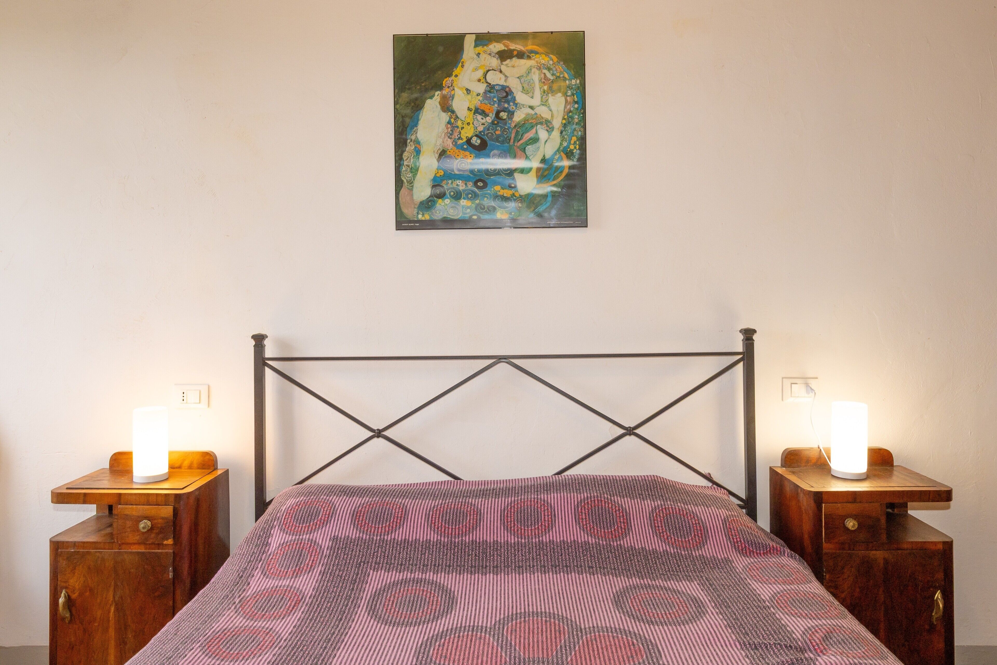 A bedroom with a bed, two bedside tables with lamps, a wall-mounted painting, and a wooden headboard.