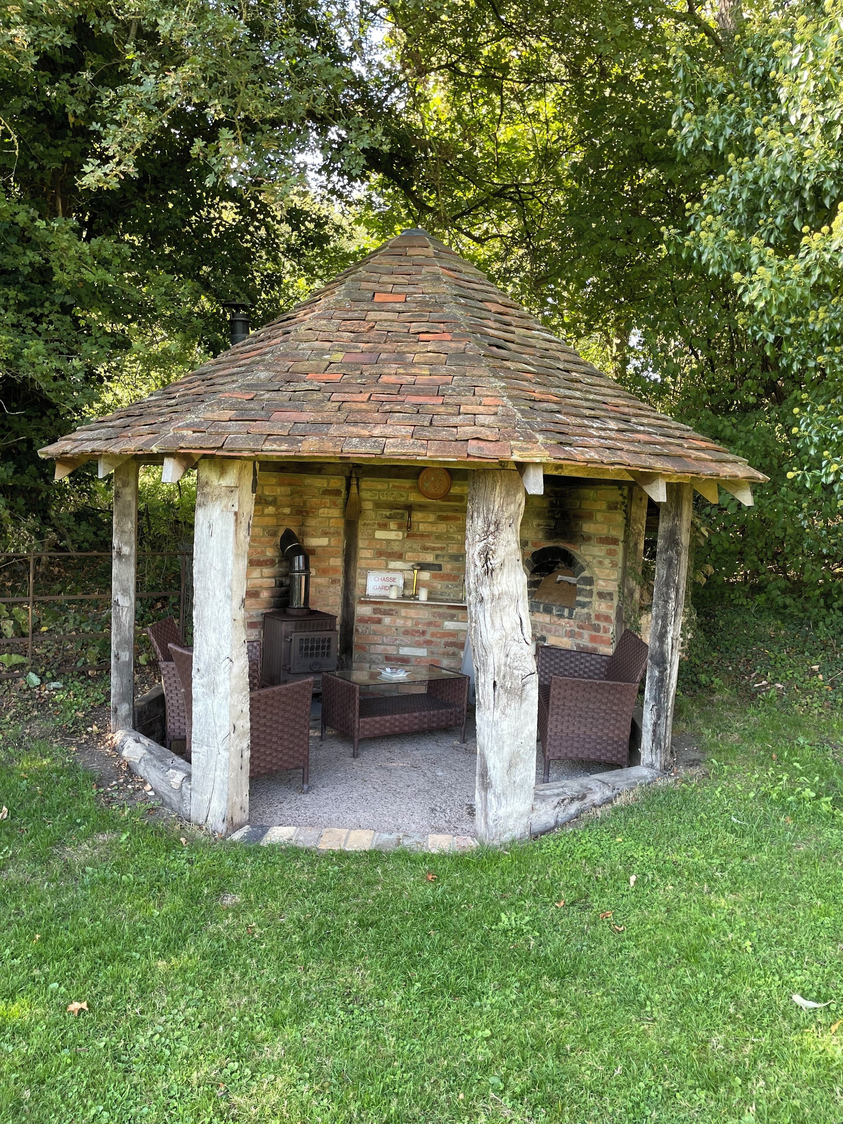 A gazebo with a brick roof and wooden pillars, featuring a fireplace and seating area.