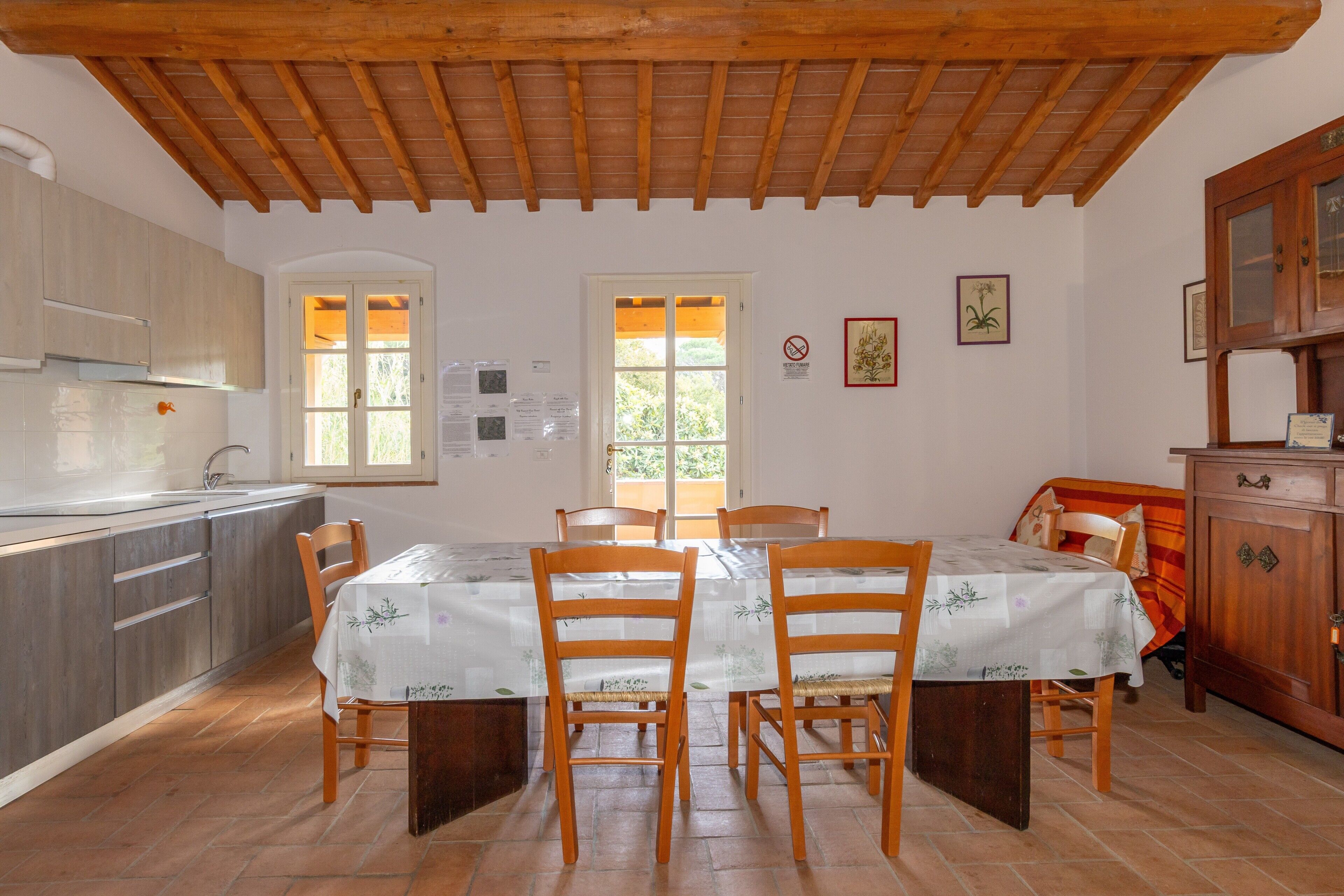 A dining area with wooden chairs and a table, a kitchen with cabinets, a wooden ceiling, and a window with a view of greenery.