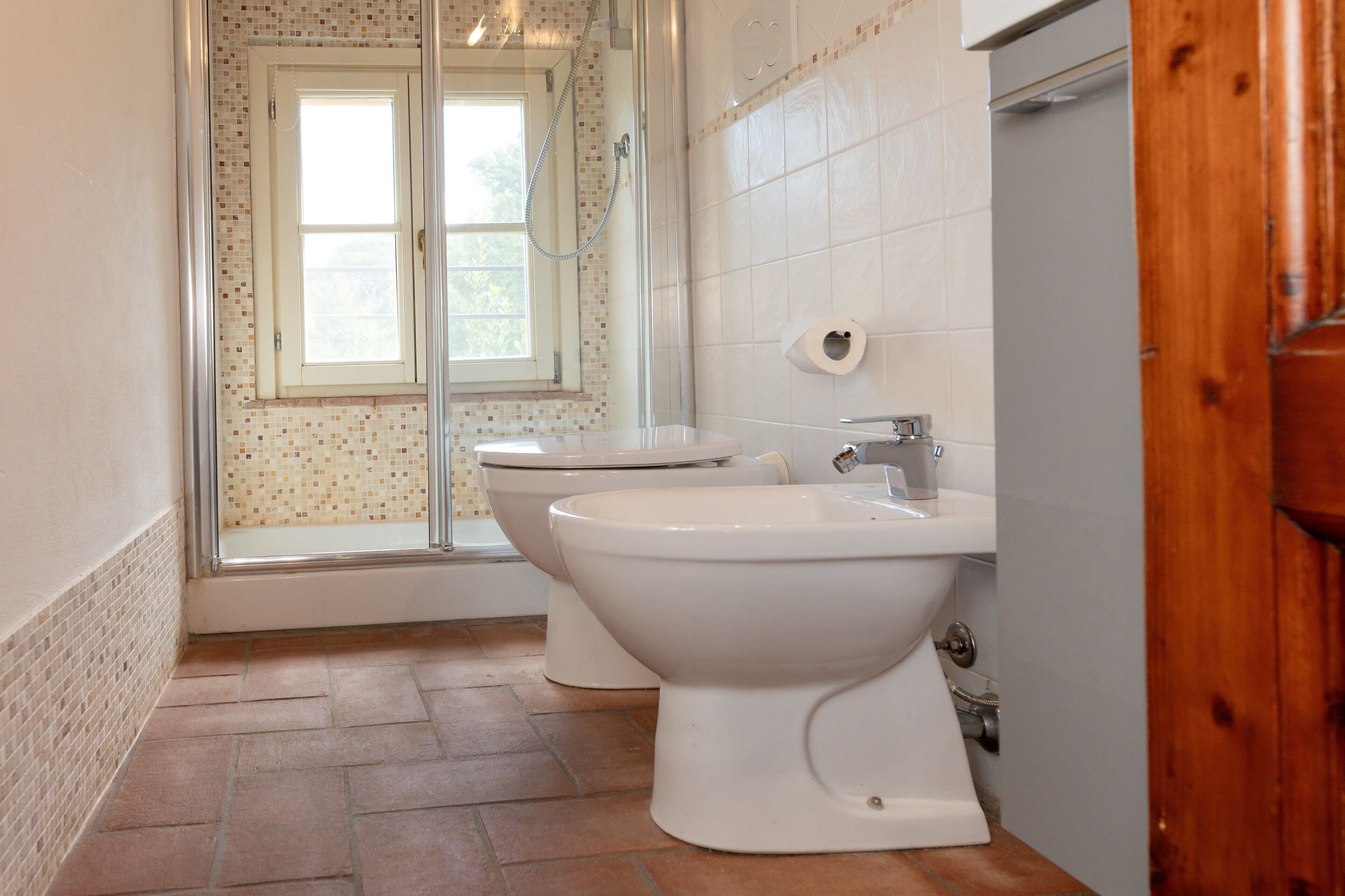 A bathroom with two toilets, a shower, and a window.