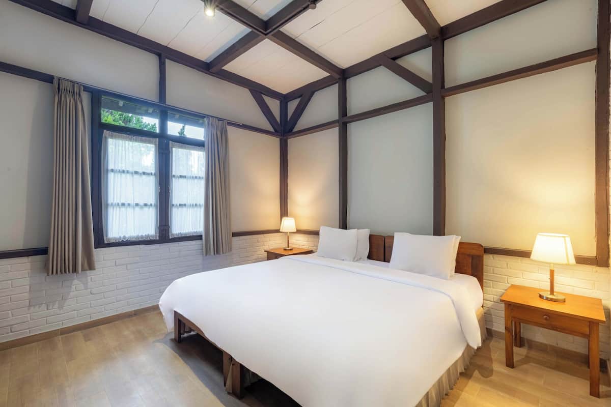 Quarto com uma cama grande, duas mesas de cabeceira com abajures, uma janela com cortinas e vigas de madeira expostas no teto.