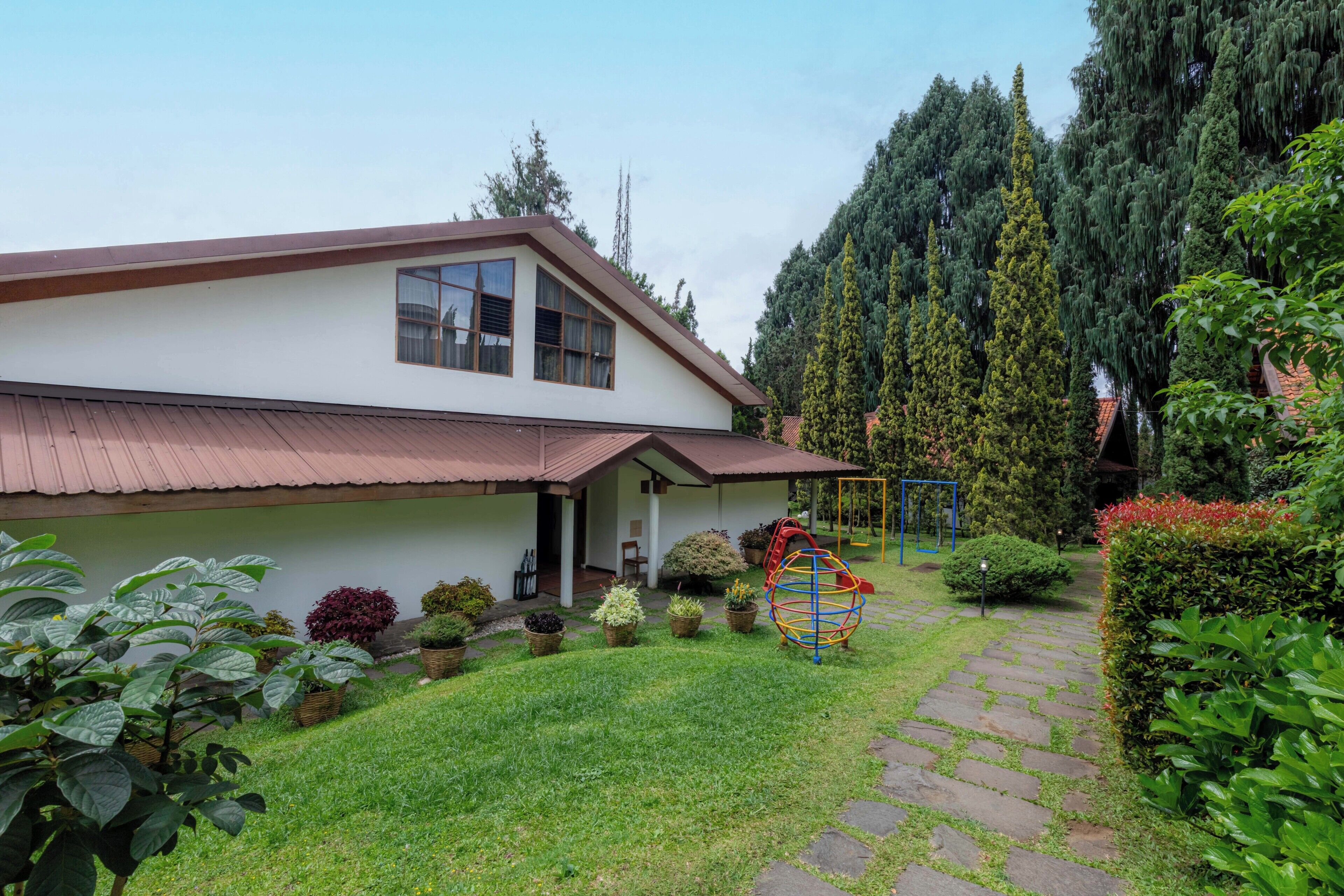 Une maison à deux étages avec un toit rouge, un porche à l’avant et un jardin où poussent différentes plantes, ainsi qu’un ensemble de jeux avec une balançoire.