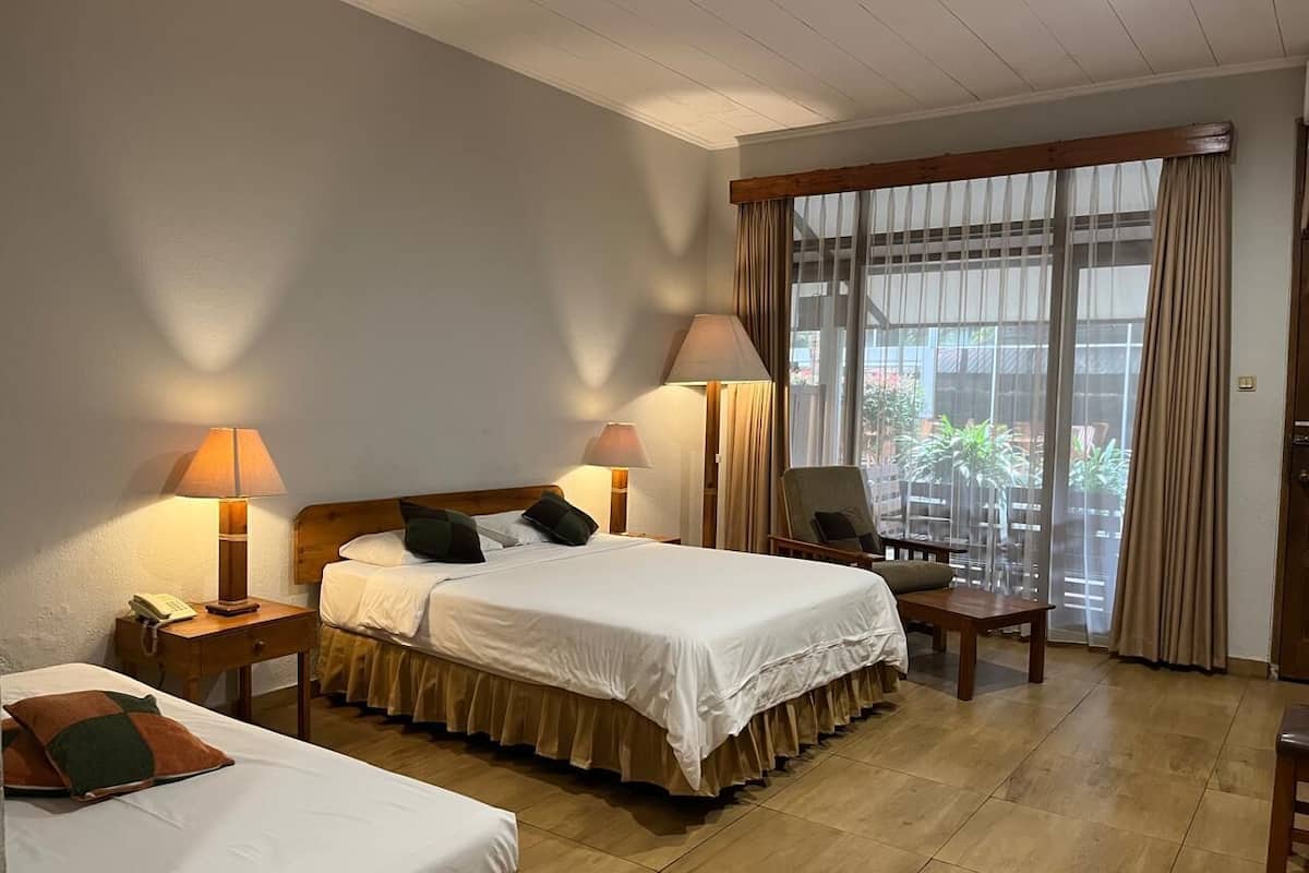 Quarto de hotel com duas camas, uma escrivaninha de madeira, uma cadeira e uma janela grande com cortinas.