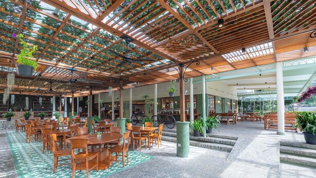 An indoor patio area with wooden tables and chairs, potted plants, and a pergola with climbing plants. - Grand Panorama Lembang (Lembang)