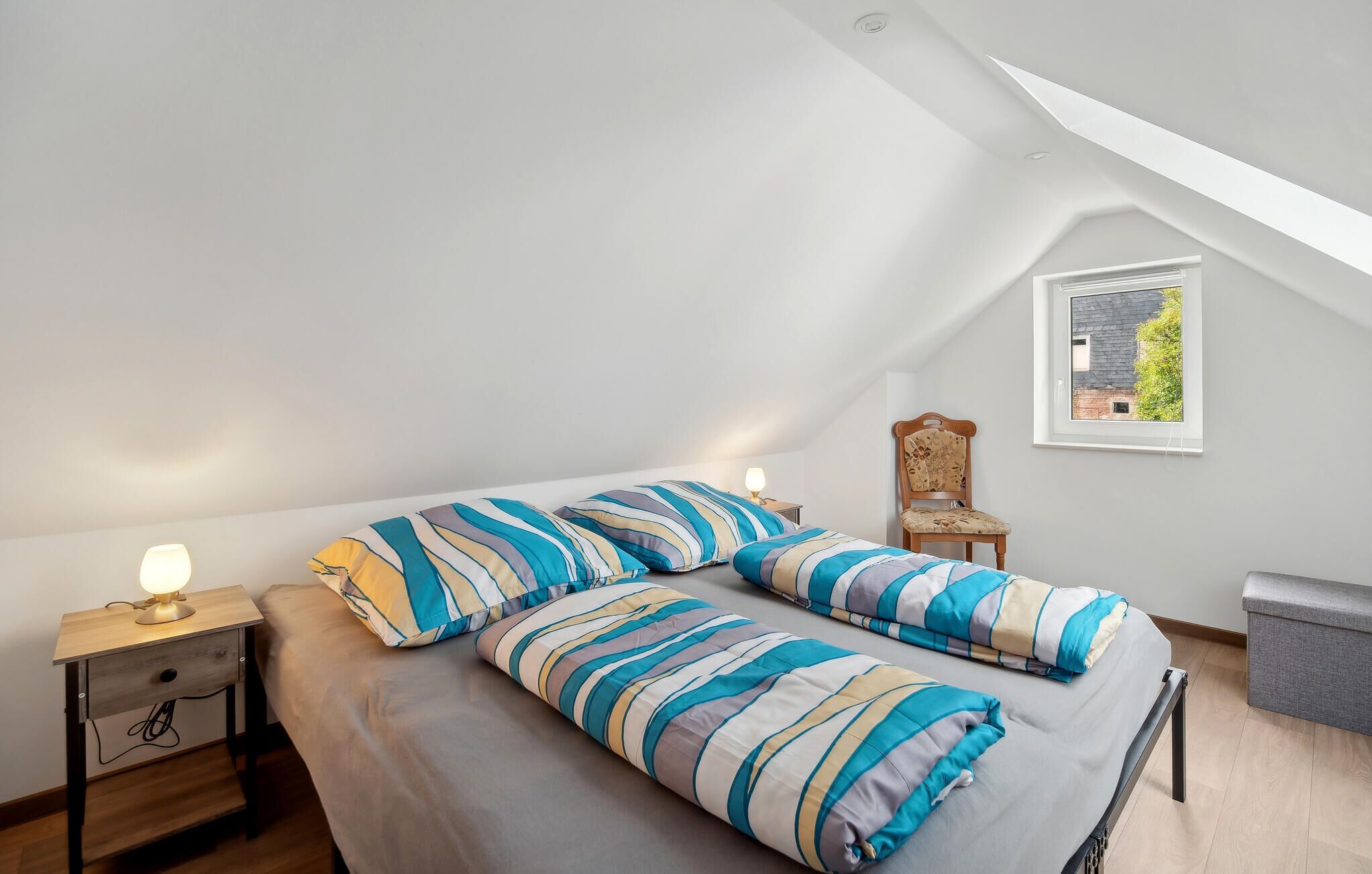 A bedroom with a sloped ceiling, a bed with striped bedding, a nightstand with a lamp, a chair, and a window with a view of greenery.