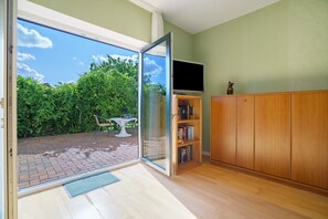 Une pièce dotée d’une porte-fenêtre coulissante donnant sur une terrasse en briques, d’un meuble en bois et d’un téléviseur fixé au mur.
