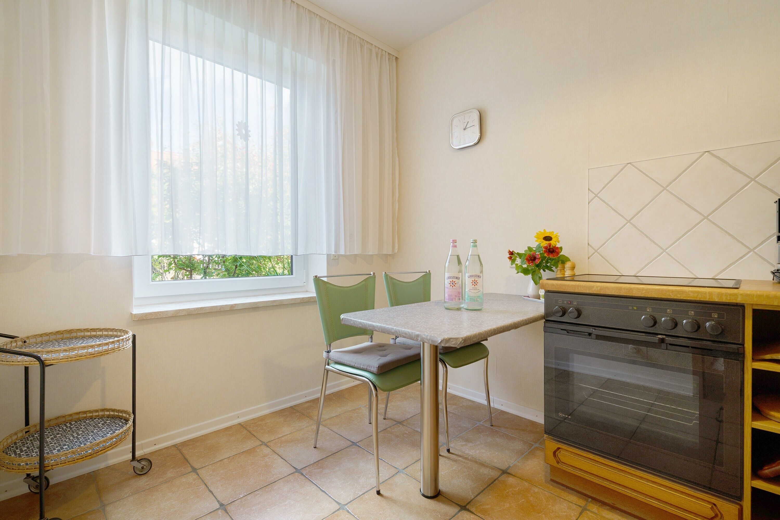 Una cocina con ventana, estufa, mesa con sillas y un carrito pequeño.
