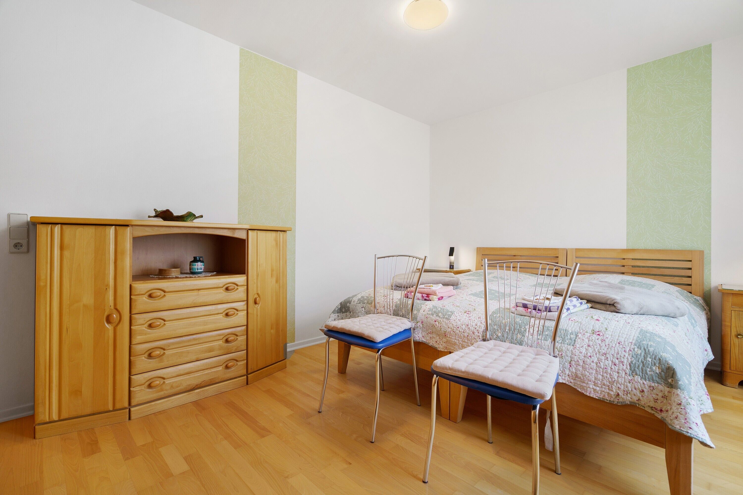 Une chambre avec une armoire en bois, deux chaises, un lit recouvert d’une couvre-lit à motifs floraux et une petite table de chevet.