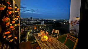A balcony with a table and chairs, decorated with flowers and a candle, overlooking a cityscape at night.