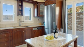 A kitchen with wooden cabinets, a stainless steel refrigerator, a granite countertop, and a bowl of grapes. - Country House 'Faith - Ios' with Sea View, Wi-Fi and Air Conditioning (ΙΟΣ)