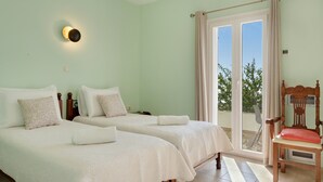 A room with two beds, a chair, a wall-mounted light, and a sliding glass door to the outside. - Country House 'Faith - Ios' with Sea View, Wi-Fi and Air Conditioning (ΙΟΣ)