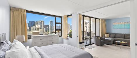 A modern hotel room with a large bed, a desk, a sofa, and a view of the cityscape.