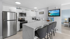 A modern kitchen with white cabinets, stainless steel appliances, a marble island with bar stools, and a flat-screen TV mounted on the wall. - Waikiki Banyan 1BR (Honolulu)