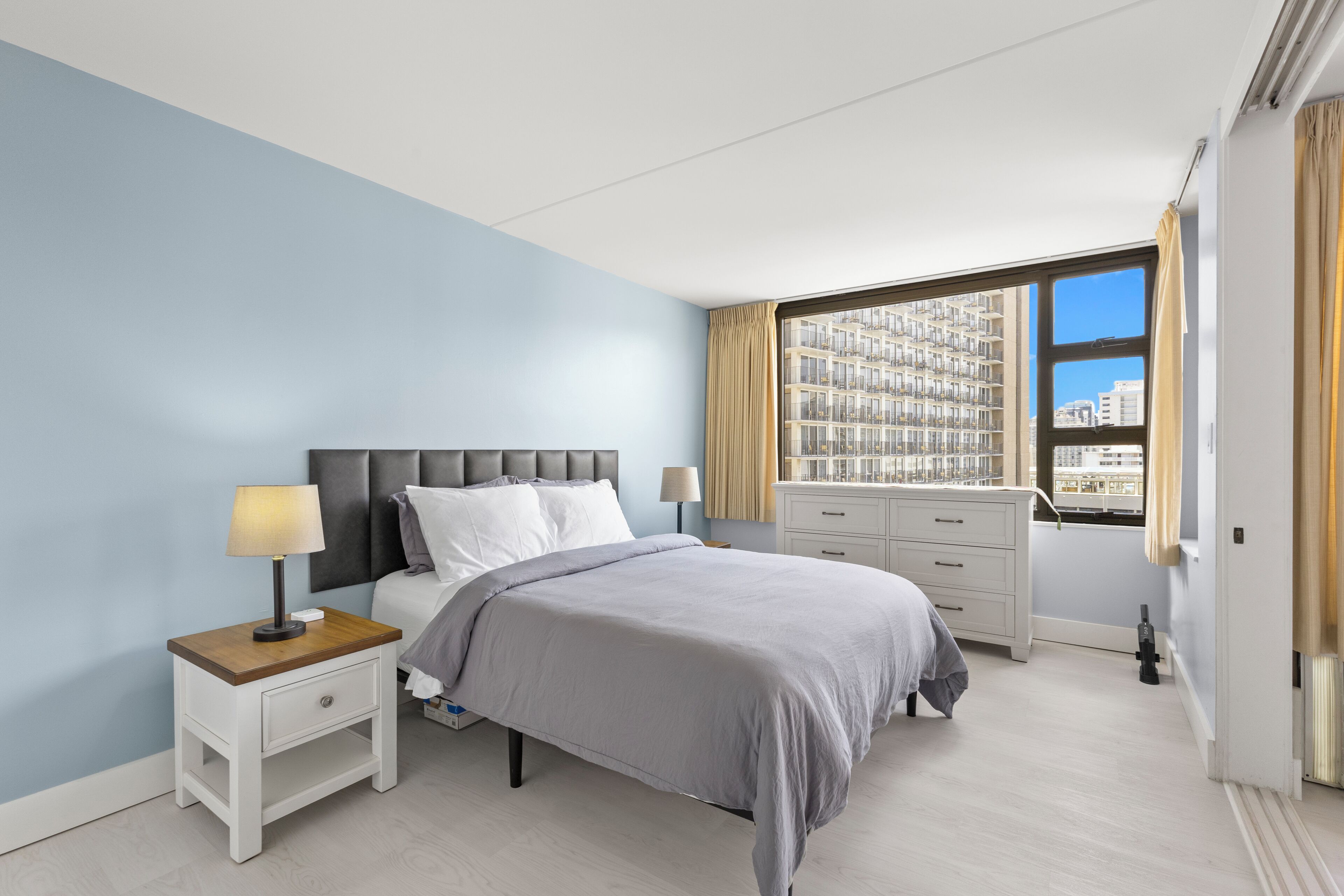 Une chambre moderne avec un lit, des tables de chevet, une commode et une grande fenêtre donnant sur la ville.