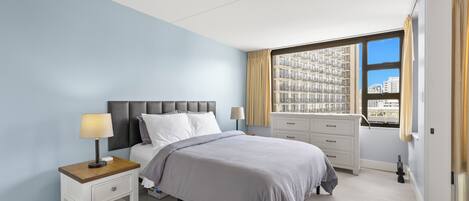 Une chambre moderne avec un lit, des tables de chevet, une commode et une grande fenêtre donnant sur la ville.