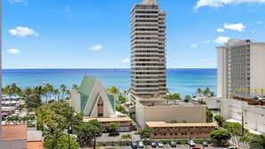 A tall hotel with a modern design, located near a beach with palm trees and a clear blue sky. - Waikiki Banyan 1BR (Honolulu)