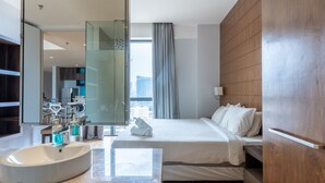 A modern hotel room with a large bed, a built-in vanity with a white sink, a view of the city through a large window, and a wooden headboard.