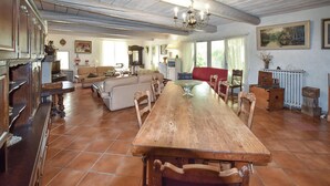 Ein geräumiger Essbereich mit einem langen Holztisch und Stühlen, umgeben von einer Küche und einem Wohnbereich mit traditioneller Einrichtung und einem großen Fenster.